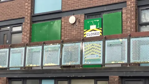 Photograph of a home on the Mottram Street estate, which has been closed by the landlord. Windows and doors are covered with green boards and there is a large sign which reads 'closed'. The balcony is metal and partially rusted. 