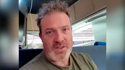 A screengrab from a video call showing a middle aged man with a short beard and short hair looking into the camera, wearing a green t-shirt. He is inside a caravan. 