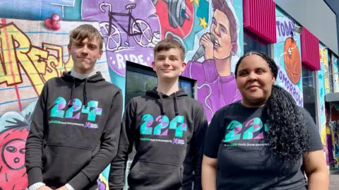 Three young people wearing black hoodies which have '224' printed on them in green and purple. They are standing next to each other smiling at the camera, with a colourful mural behind them. 