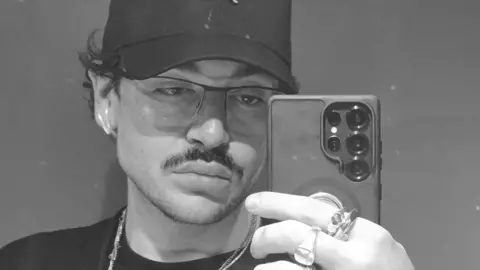 Paul Aaron Paul Aaron, who is wearing a baseball hat and glasses, is taking a selfie in a mirror. 