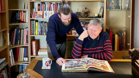 Karl Hansen Peter Hansen and his son Karl. They are looking down at a large hardback comic book, lying open on a desk.
