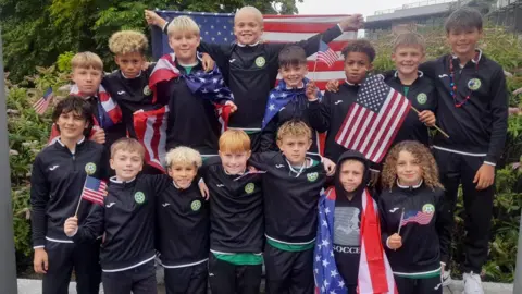 a group of young footballers pose in team colours. Some are holding US flags