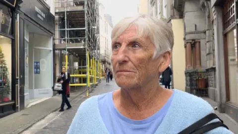 Cathy has short white hair. She is wearing a purple t-shirt with a baby blue v-neck jumper on top.