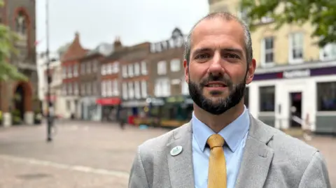 Stuart Gourley is stood in Newbury town centre, wearing a light blue shirt and yellow tie.