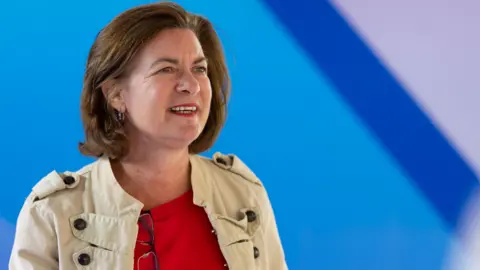Getty Images Eluned Morgan - A woman with brown hair looking to the left of the camera. She is wearing a red top and a beige jacket. There is a blue background
