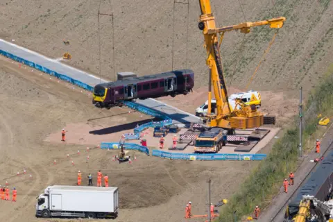 Bav Media A giant yellow crane in a field next to railway line. It is lifting a train carriage. A temporary track has been built to get the crane into position in the field. There are about a dozen people in orange overalls.