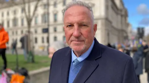 A man wearing a suit looks into the camera. He has short, grey hair, swept back and wears a dark blue suit over a blue jumper and shirt. Behind him is a big stone, beige-coloured building.