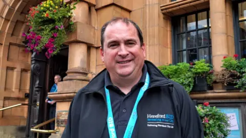 A man wearing dark clothing and a light blue lanyard is outside a building. He has dark hair and is looking at the camera.