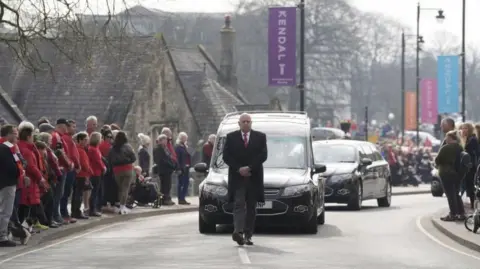 A man walks ahead of a funeral cortege on an otherwise empty road with wide pavements. It is lined with people many of them wearing red. 
