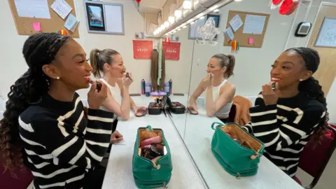 Shaun Whitmore/BBC Sydney Wimes at the front is in a white and black striped jumper and is looking in the mirror applying red lipstick. In the background is another performer who is looking into the mirror and applying makeup to her face.