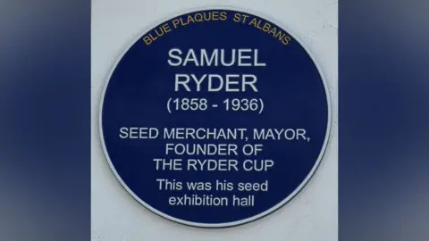 Snjezana Boatswain The image shows a round blue commemorative plaque mounted on a light-coloured wall. The plaque honours Samuel Ryder, a British businessman and civic leader best known for founding the Ryder Cup golf tournament. It indicates that the building where the plaque is installed was once his exhibition hall.