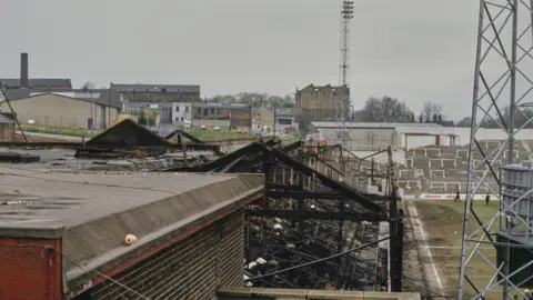 Getty Images The fire-damaged skeleton of a football stand at Bradford City's Valley Parade ground, pictured in 1985. Another terraced stand, undamaged, can be seen in the distance, with an electric pylon in the foreground.