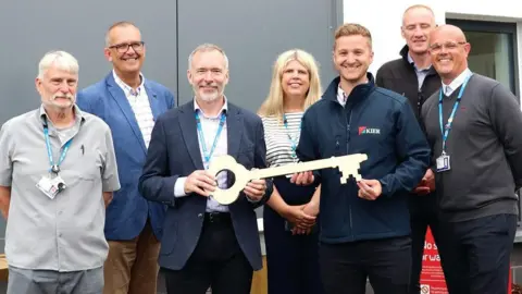 Dr Francis Old, Chris Couch, Steve Williamson, Lynda Sherfield-Wong, Karl Martin, Jeff Houghton and Lewis Saundry are standing in a line smiling outside the building. Mr Williamson is holding a giant key.