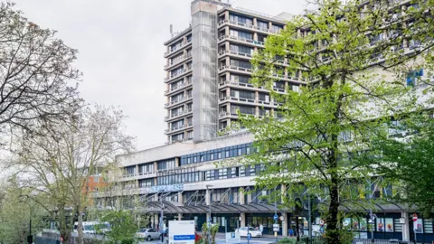 Exterior of Royal Free Hospital in north London, showing a glass awning outside the building, a car park and a vertical white street sign for the hospital