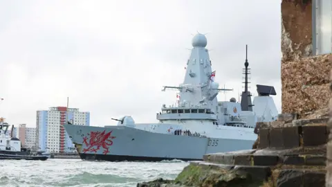 HMS Dragon seen departing Portsmouth Harbour with rocks in the foreground and building behind. We can see the front half of the ship. The red dragon logo on the ship is clearly seen 