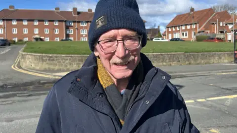 Norman Dennis stands on the pavement at the side of the road. He is wearing a navy woolly hat, a large navy jacket and a yellow and black scarf. He also wears glasses, has a moustache and is smiling at the camera. 