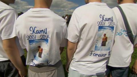 The backs of four boys, two centred, wearing t-shirts with tributes to a teenager who died from a falling tree in Carlton-in-Lindrick, Nottinghamshire
