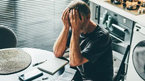 Getty Images A man sits with his face in his hands at a kitchen table, receipts and a notebook in front of him. An oven and washing machine are behind him and a window with his blinds to the right. He is wearing a dark grey t-shirt. His hair is grey.