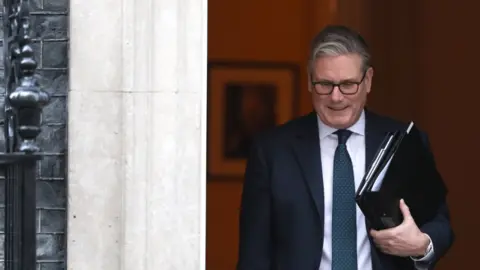 Prime Minister Keir Starmer departs Downing Street ahead of his statement on Iran. He wears a black suit, white shirt and navy tie and is carrying two black folders of documents