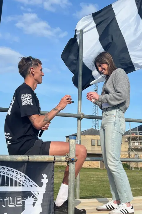 Leodhais Macpherson proposed to his girlfriend, Jaimie Walker. He is sitting down and holding a box containing a ring in his right hand. His girlfriend is smiling. She is standing and has long dark hair. She is wearing a grey jumper and pale blue jeans with white trainers.