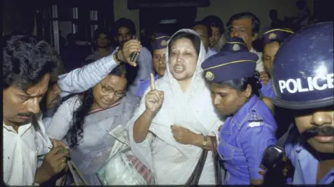 Getty Images Khaleda Zia is wearing a white headscarf and saree and gesturing with a raised finger, while she is taken away in a crowd by a group of police officers in blue uniforms and hats
