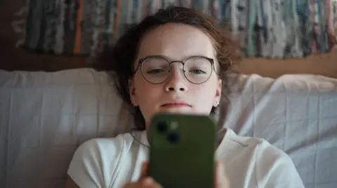 A young teenage girl looks at a smartphone. She is wearing glasses and a white T-shirt and leaning against white pillows or cushions with coloured curtains in the background.