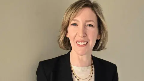 Dr Gillian Brooks A woman is smiling at the camera, wearing subtle makeup. She has short blonde  hair and is wearing a black blazer with a detailed gold necklace. 
