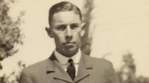 Great Ayton History Society A vintage black‑and‑white portrait of a person in a formal suit and tie, standing outdoors with blurred trees in the background.
