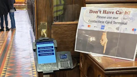 An electronic tablet is fixed to a stand at the inside door of a church. To the left, legs and feet can be seen standing up a tiled aisle. To the right, on top of wooden unit there is a sign that says: "Don't have cash? Please use our contactless card terminal." 