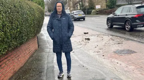 Sarah Dewart A woman with blue hair and black spectacles wearing a blue padded coat standing on an asphalt and brick pavement. A pool of water with excrement and paper flows down the path in the right of the picture.
