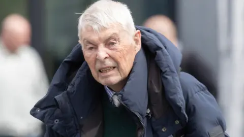 A man with short grey hair is looking into the distance to the left of the camera. He is wearing a large navy coat with a hood draped behind his head. The man is dressed in a pinstripe shirt and tie. The background to the image is out of focus and shows two other men in the distance. 