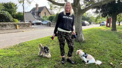 Kevin Shoesmith/BBC Simone Hunt, with blonde shoulder-length hair, wearing a black long-sleeved top with a white Adidas logo and green and brown camouflaged trousers, pictured with two small dogs on leads. 