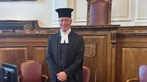 Guernsey's retiring HM Greffier Jon Torode wearing his ceremonial black robe and hat. He is standing inside a courtroom at the Royal Court of Guernsey. In front of him is the a table which has a computer on it, a microphone, mini TV screen and a digital clock.