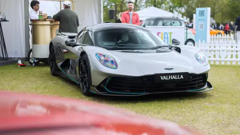 Getty Images A silver grey Aston Martin Valhalla on grass, at an event. There arethree people in hte background