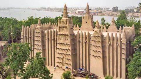AFP via Getty Images The giant mud structure of the Grand Mosque of Mopti surrounded by trees, with a river seen in the background.