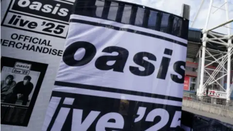 Large black and white banners outside a venue. One says Oasis Live 25, the other says Oasis Live 25 official merchandise