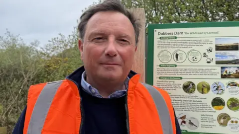 Brown haired man wearing a high viz jacket and a blue sweater is standing next to an information board about dubbers and the wildlife you might see at the location