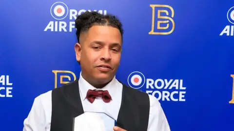 Ben Butler A man with dark curly hair, a white shirt, red bow tie and black waistcoat with something shiny in his hands and a blue background with Royal Air Force written on it