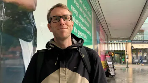 A white male wearing dark-framed glasses and a black and cream rain coat looks at the camera