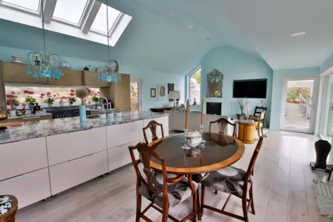 IWC Media A bright, open-plan kitchen and dining space features pale blue walls, skylights, and large windows that flood the room with natural light.
A marble-topped island, modern cabinetry, and a round wooden dining table with chairs create a blend of contemporary and classic design.