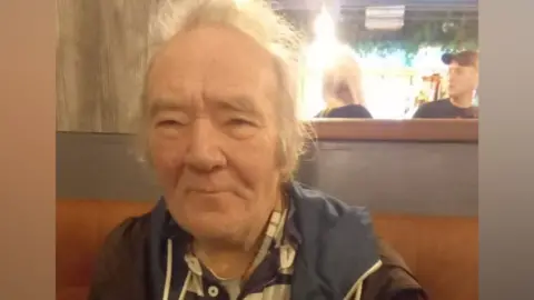 PSNI/FAMILY HANDOUT An older man with receding white hair. He is wearing a blue and brown jacket with a blue and brown shirt underneath