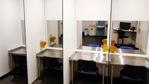 Interior of consumption room, showing four individual desks and chairs with a mirror, separated by divider screens. Each area contains a sharps box.