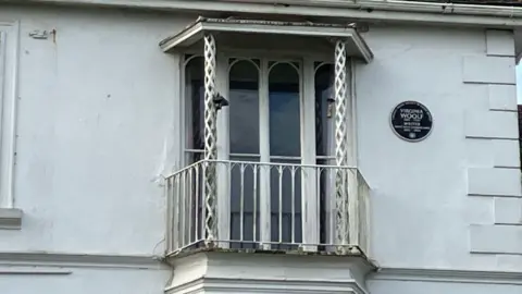 Exterior de um conjunto de janelas francesas com varanda. Uma placa preta em homenagem a Virginia Woolf está fixada na parede à direita dela.