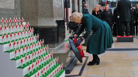 PA Media Queen Camilla bends down to place her wreath at a memorial in Paddington station. It is surrounded by a display of small wooden crosses with poppies on. Behind her, a choir is singing.