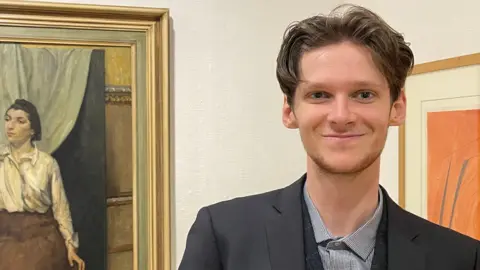 A young man smiles at the camera. He wears a smart suit and stands in front of works of art in a gallery.