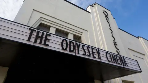 An exterior shot of The Odyssey Cinema in St Albans with the wording of the cinema on the outside of a white art deco building, taken on a clear day with blue skies.