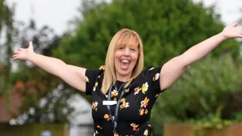 Tracey Richardson Woman smiling with hands in the sky with a black dress with flowers on it.