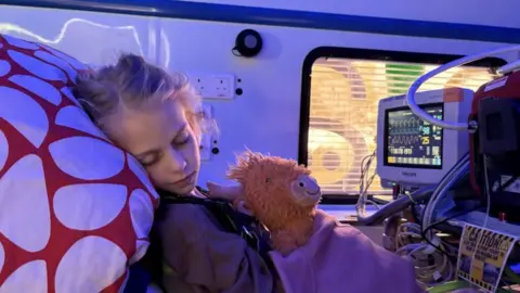 Air Ambulance Service A young girl appears to be sleeping while travelling in an ambulance. She is in a hospital and is resting on a pillow while surrounded by medical equipment. She is holding a cuddly soft toy of a cow.