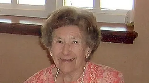 A brown-haired woman smiling. She is wearing a pink top, pearl necklace and pearl earrings