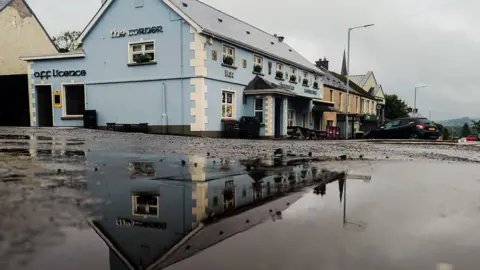 Aisling Kelly A large puddle on the ground outside a blue building. The building is in the reflection of the puddle.